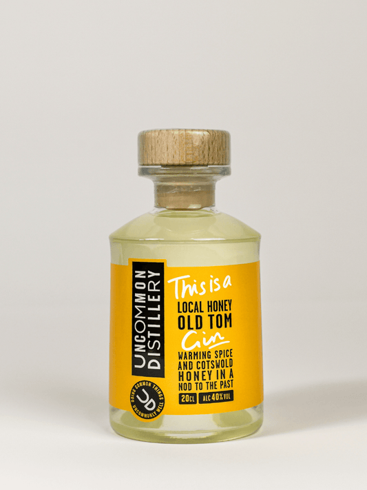 A clear glass bottle of Uncommon Distillery 'This is a Local Honey Old Tom' spice and citrus honey blend, featuring a bright yellow label and a light wood-grain cap, set against a neutral light gray background.