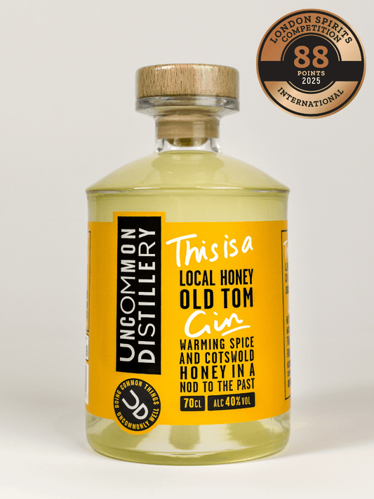 A bottle of Uncommon Distillery 'This is a Local Honey Old Tom Gin' with a yellow label and a wooden cap, shown on a light gray background with a London Spirit's Competition 2023 award badge in the top right corner.