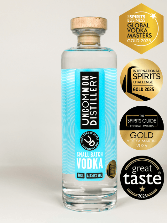 A clear glass bottle of Uncommon Distillery Small Batch Vodka with a light wood cap and a bright turquoise label, shown on a light gray background with four gold and black award badges on the right.