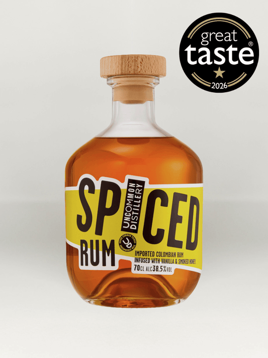 A bottle of Uncommon Distillery Spiced Rum with amber liquid, a light wood cap, and a yellow and black label, shown on a light gray background with a Great Taste 2026 award badge in the top right corner.
