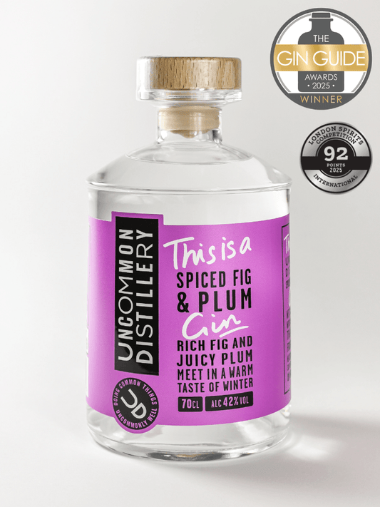 A 70cl clear glass bottle of Uncommon Distillery 'This is a Spiced Fig & Plum Gin' with a purple label and a light wood cork stopper. The bottle is set against a light gray background with two award badges in the top right corner: 'The Gin Guide Awards 2023 Winner' and 'London Spirit Awards 2023 92nd Position'.
